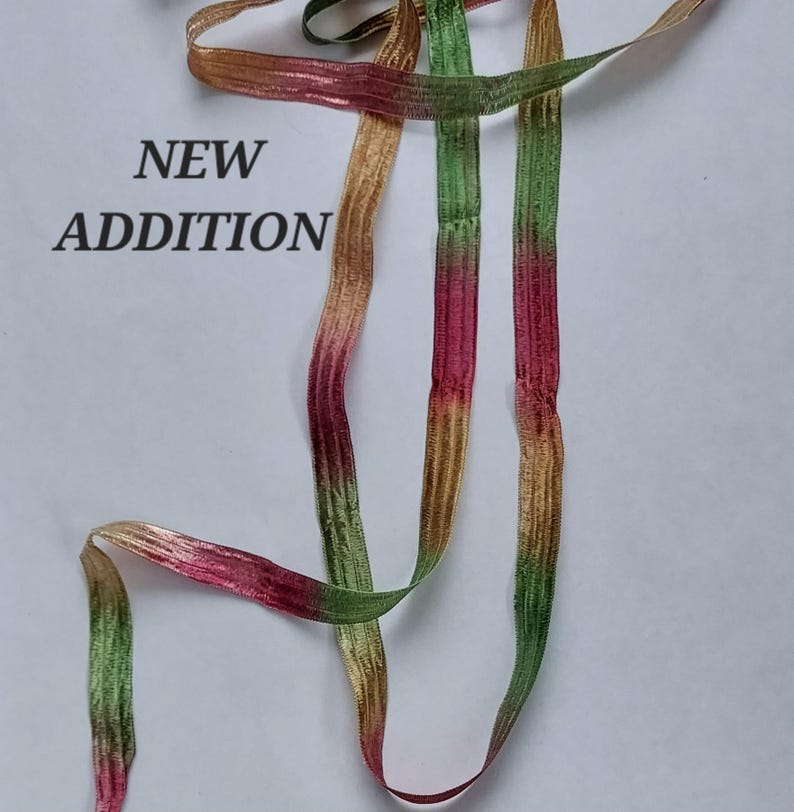 May include: A length of ribbon featuring a gradient color pattern of green, pink, and gold. The ribbon has a textured, woven appearance. The text NEW ADDITION is printed in black on the left side of the image, next to the ribbon strands.