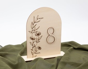 May include: Wooden table number with a floral design and the number 8. The number is cut out of the wood and the design is engraved. The table number is on a green fabric surface.