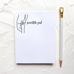 May include: A white notepad with a black line drawing of a person scratching their back. The notepad has the text "scratch pad" written on it.