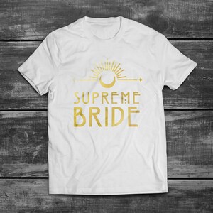 May include: White t-shirt with a gold graphic of a sun, moon, and the text "SUPREME BRIDE".