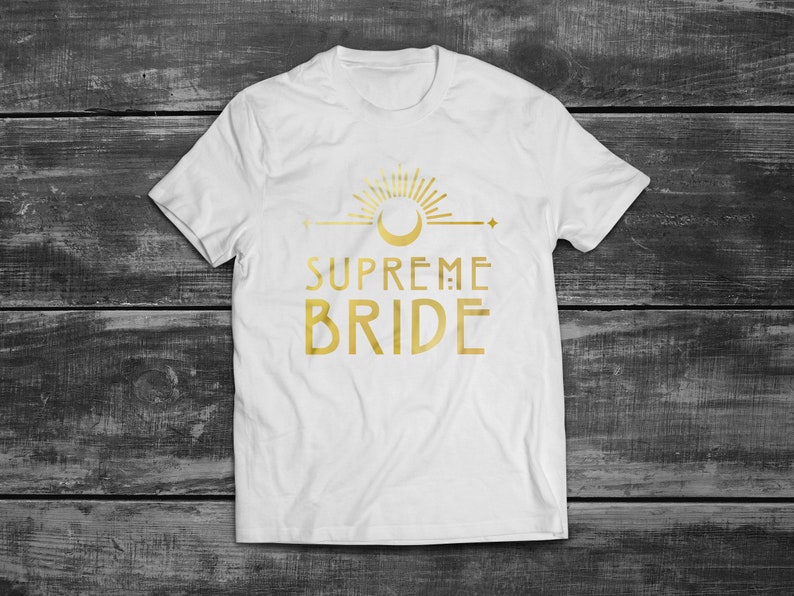 May include: White t-shirt with a gold graphic of a sun, moon, and the text "SUPREME BRIDE".