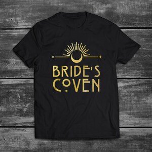 May include: Black t-shirt with a gold graphic design featuring a crescent moon, sun, and stars. The text "Bride's Coven" is written in a stylized font.