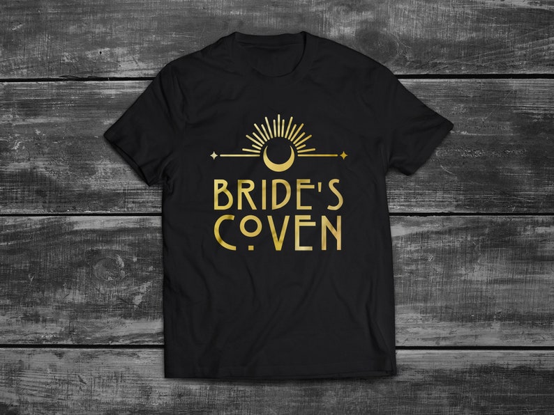 May include: Black t-shirt with a gold graphic design featuring a crescent moon, sun, and stars. The text "Bride's Coven" is written in a stylized font.