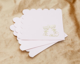 May include: A stack of pink paper napkins with scalloped edges. The napkins feature a gold foil design of two champagne glasses clinking together with the word "Cheers!" printed below.