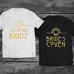 May include: Two white and black t-shirts with gold foil designs. The white shirt says "Supreme Bride" with a crescent moon and sunburst design. The black shirt says "Bride's Coven" with a crescent moon and sunburst design.