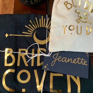 May include: A black t-shirt with gold foil lettering that reads "Bride Coven". A small white bag with gold foil lettering that reads "But Did You Die?" and a skull and crossbones. A black tag with white lettering that reads "Jeanette".