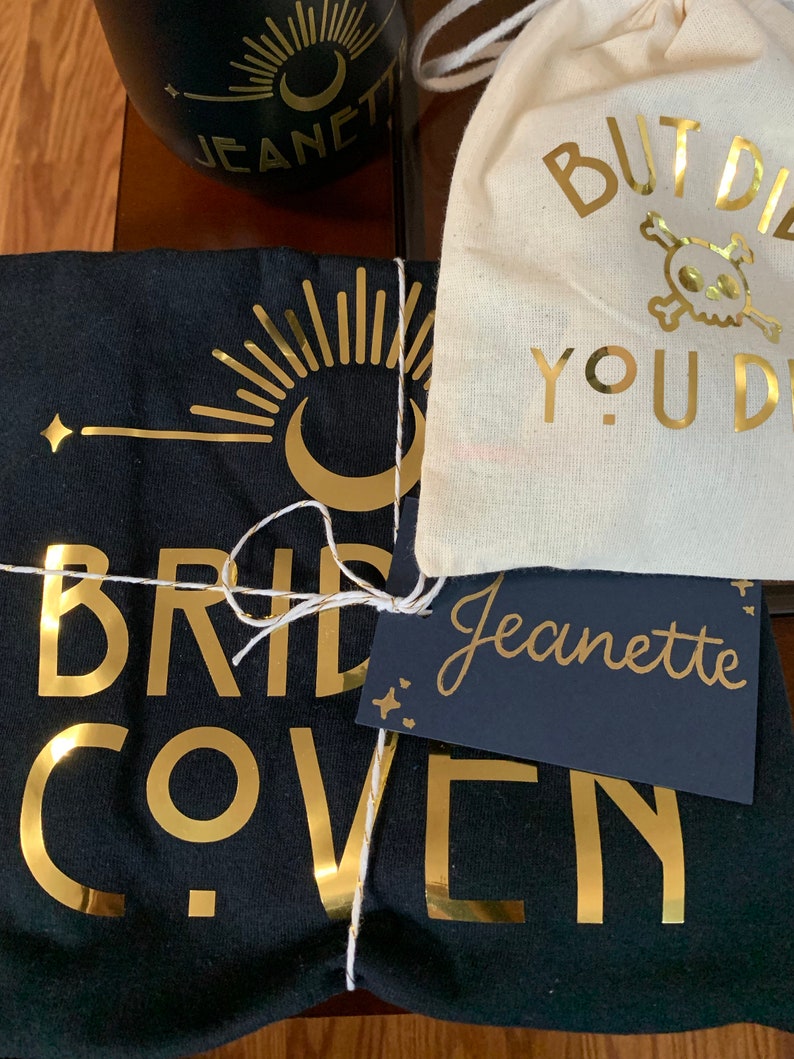 May include: A black t-shirt with gold foil lettering that reads "Bride Coven". A small white bag with gold foil lettering that reads "But Did You Die?" and a skull and crossbones. A black tag with white lettering that reads "Jeanette".