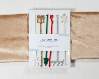 May include: A set of six Christmas cocktail stir sticks in a clear plastic package. The sticks are decorated with glitter and feature various holiday designs, including a snowflake, a Christmas tree, a present, a candy cane, a snowman, and a snowflake.