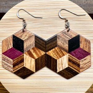 May include: Geometric wooden earrings with a mosaic design. Each earring is a hexagon shape with a 3D cube pattern in various wood tones, including black, light brown, and deep purple. The earrings hang from silver-colored fish hook ear wires.