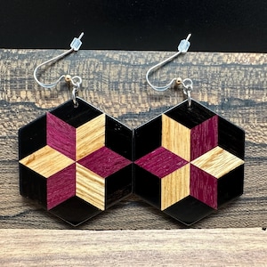 May include: A pair of hexagonal earrings featuring a geometric star pattern composed of inlaid wood segments in black, deep purple, and light tan. The earrings hang from silver-toned metal fishhook wires with small clear plastic backings.