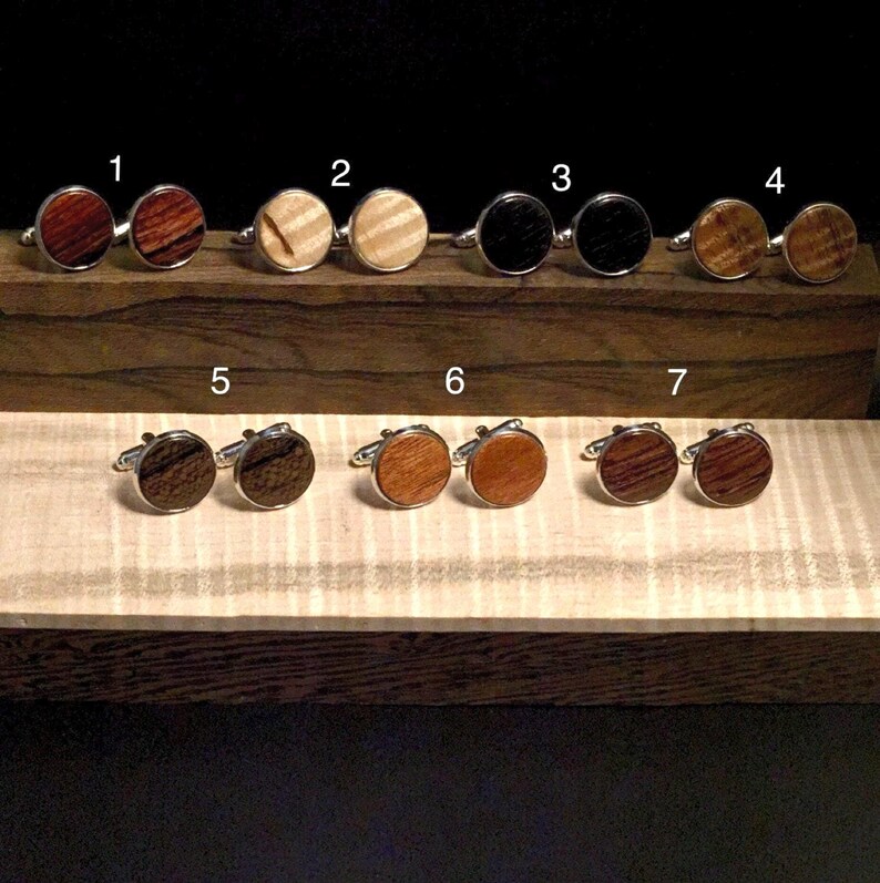 K&ouml;nnte beinhalten: Eine Sammlung von sieben Paaren runder Manschettenkn&ouml;pfe aus Holz mit silberfarbenen Metallr&uuml;cken. Die Manschettenkn&ouml;pfe weisen verschiedene Holzmaserungen und -farben auf, darunter Dunkelbraun, Hellbraun und Schwarz. Die Manschettenkn&ouml;pfe werden auf einer Holzoberfl&auml;che pr&auml;sentiert.