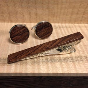 K&ouml;nnte beinhalten: Ein Set aus Manschettenkn&ouml;pfen und einer Krawattenklammer aus Holz. Die Manschettenkn&ouml;pfe sind rund mit silbernem Rand und einem dunkelbraunen Holzmaserungszentrum. Die Krawattenklammer ist rechteckig, aus dem gleichen Holz und hat eine silberne Klammer.