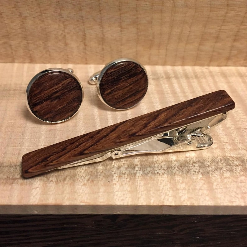 K&ouml;nnte beinhalten: Ein Set aus Manschettenkn&ouml;pfen und einer Krawattenklammer aus Holz. Die Manschettenkn&ouml;pfe sind rund mit silbernem Rand und einem dunkelbraunen Holzmaserungszentrum. Die Krawattenklammer ist rechteckig, aus dem gleichen Holz und hat eine silberne Klammer.