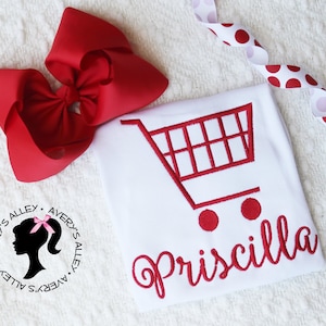 May include: White t-shirt with a red shopping cart graphic and the name Priscilla in red cursive lettering. The t-shirt has a small black silhouette of a woman with a pink bow in her hair and the text Avery's Alley repeated around her.