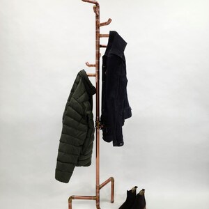 Puede incluir: Un perchero de tuber&iacute;a de cobre con m&uacute;ltiples ganchos, sosteniendo una chaqueta acolchada verde oscuro y una chaqueta negra. Botines negros en la base. El perchero tiene un dise&ntilde;o industrial moderno.