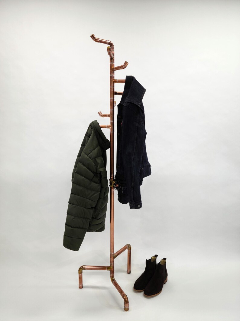 Puede incluir: Un perchero de tuber&iacute;a de cobre con m&uacute;ltiples ganchos, sosteniendo una chaqueta acolchada verde oscuro y una chaqueta negra. Botines negros en la base. El perchero tiene un dise&ntilde;o industrial moderno.