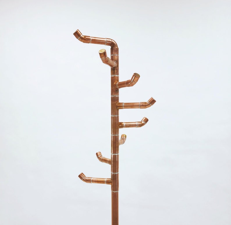 Puede incluir: Un perchero de color cobre con un dise&ntilde;o en forma de &aacute;rbol. El perchero presenta m&uacute;ltiples brazos curvos que se extienden desde un poste central, proporcionando espacio para colgar prendas. El acabado met&aacute;lico le da una est&eacute;tica moderna e industrial.