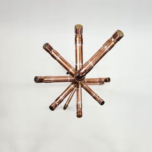 Puede incluir: Una escultura en forma de estrella de color cobre. La escultura est&aacute; hecha de tuber&iacute;as y accesorios de cobre, con una esfera central y brazos radiales. Los extremos de las tuber&iacute;as est&aacute;n cubiertos con un metal m&aacute;s oscuro.