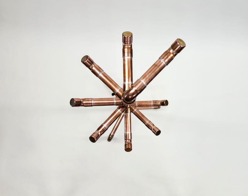 Puede incluir: Una escultura en forma de estrella de color cobre. La escultura est&aacute; hecha de tuber&iacute;as y accesorios de cobre, con una esfera central y brazos radiales. Los extremos de las tuber&iacute;as est&aacute;n cubiertos con un metal m&aacute;s oscuro.