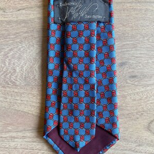 May include: A blue silk tie with a red and white geometric pattern. The tie has a burgundy lining and a black label that reads "Exclusively for Jaffe Kensington".