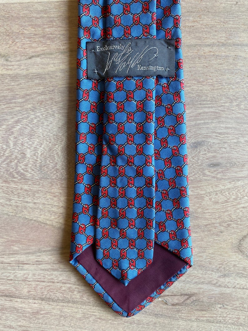 May include: A blue silk tie with a red and white geometric pattern. The tie has a burgundy lining and a black label that reads "Exclusively for Jaffe Kensington".