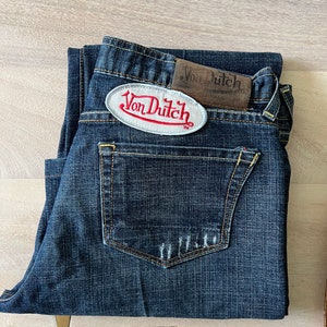 May include: A pair of dark blue denim jeans with a white patch that says "Von Dutch" in red lettering. The jeans have a back pocket with a brown leather patch that says "Von Dutch" in white lettering.