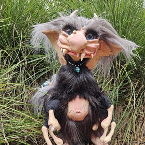 May include: A handmade troll doll with gray hair, a pink face, and large blue eyes. The troll is wearing a black shirt and has a black fur collar with a blue skull charm.