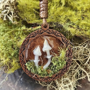 May include: A circular wooden pendant featuring two small white mushroom figures encased in a clear resin layer. The wooden frame is engraved with a continuous black knotwork pattern. The pendant hangs from a brown cord with a knotted loop.