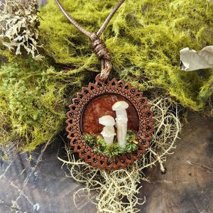 May include: A circular pendant featuring two small, translucent white mushroom figures set against a dark reddish-brown background, encased in a decorative brown wooden frame with a scalloped edge, suspended from a thin brown cord.