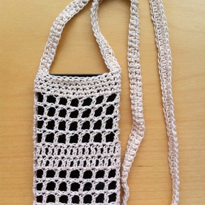 Puede incluir: Una funda de teléfono de crochet blanco con una correa larga. La funda tiene un patrón de cuadrícula y está diseñada para sostener un teléfono inteligente.