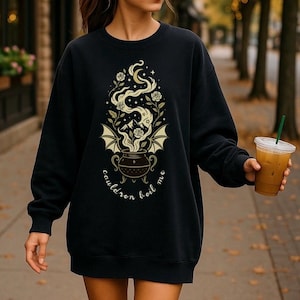 May include: Black sweatshirt with a whimsical design featuring a cauldron, roses, and bat wings, with the text "cauldron boil me". The design includes a moon and stars, creating a mystical theme. The sweatshirt is a long sleeve style.