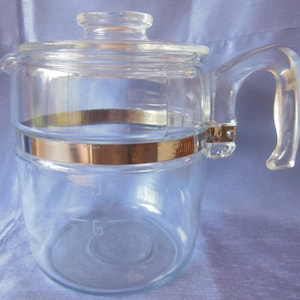 May include: A clear glass coffee pot with a lid and handle. The pot has a gold-colored band around the middle and a gold accent where the handle attaches. The pot has a capacity marking of 6.