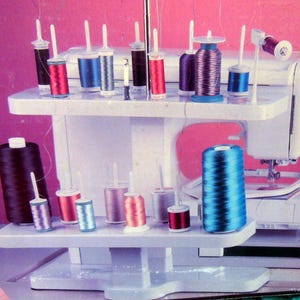May include: A white sewing machine with a thread rack holding various colored spools of thread. The machine has a needle and a small embroidery hoop. A large spool of black thread and a large spool of blue thread are also visible.