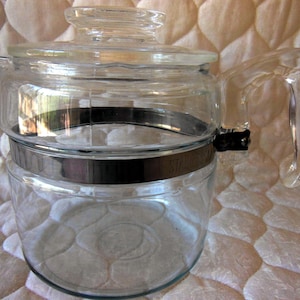May include: A clear glass teapot with a matching glass lid and a sturdy glass handle. A wide, polished stainless steel band encircles the center of the pot, featuring the embossed text "STAINLESS" on the side near the handle attachment point.