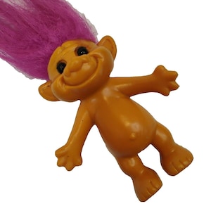 May include: A small plastic troll figurine with smooth orange skin, large ears, and a wide smiling face featuring black eyes. The figure has a prominent tuft of vibrant, long, upright magenta hair extending from the top of its head.