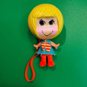 May include: A vintage doll with a yellow head and hair, a smiling face, and a blue dress. The doll has orange shoes and arms, and a red ribbon. The doll's chest has a red and yellow design.