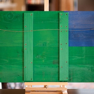 May include: A wooden sign with a green and blue paint finish. The sign has four vertical wooden strips with screws and a wire hanger attached to the top.