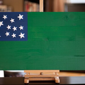 May include: A wooden flag with a blue canton and 13 white stars on a green background. The flag is displayed on a wooden easel.