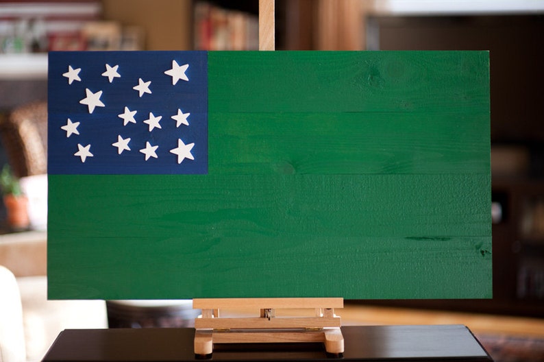 May include: A wooden flag with a blue canton and 13 white stars on a green background. The flag is displayed on a wooden easel.
