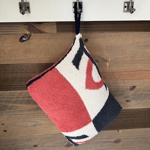 May include: A geometric patterned towel in red, white, and navy blue hangs from a hook. The towel has a loop for hanging and a textured surface. The design features abstract shapes and patterns.