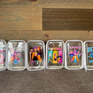 May include: Six rectangular clear plastic pencil cases with white zippers. Each case features colorful anime-style character graphics and the text "HUNTRAX" or "DERPY" printed on the front, showcasing various vibrant designs and artistic illustrations.