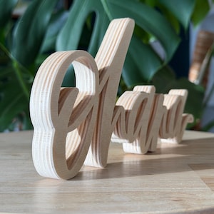 May include: Wooden sign spelling out the word "Alright" in a cursive font. The sign is made of light-colored wood and is standing on a wooden surface. The background features green foliage.