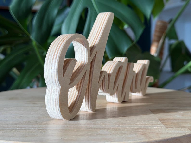 May include: Wooden sign spelling out the word "Alright" in a cursive font. The sign is made of light-colored wood and is standing on a wooden surface. The background features green foliage.