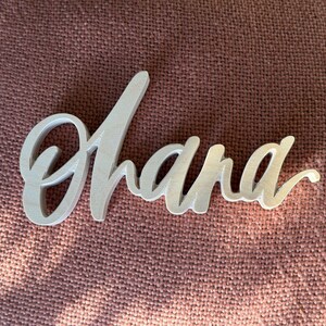 May include: A wooden sign in a cursive font that spells "Ohana". The sign is light-colored and rests on a textured, reddish-brown surface. The word is a popular Hawaiian term.