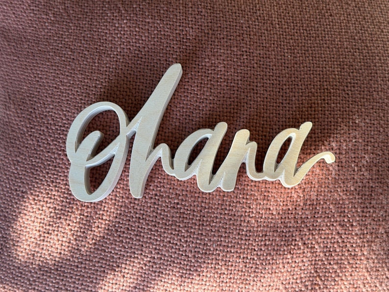 May include: A wooden sign in a cursive font that spells "Ohana". The sign is light-colored and rests on a textured, reddish-brown surface. The word is a popular Hawaiian term.