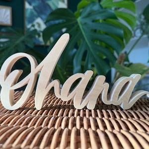 May include: A wooden sign that spells out the word "Ohana" in a cursive font. The sign is made of light-colored wood and is placed on a woven wicker surface. The background includes green plants and artwork.