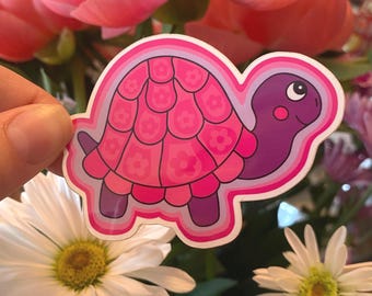 May include: A pink and purple turtle sticker with a flower design on its shell. The sticker is held in front of a bouquet of pink peonies and white daisies.