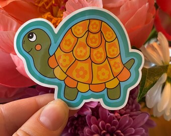 May include: A colorful turtle sticker with a retro design. The turtle has a green head and feet, an orange and yellow shell with flower patterns, and a blue outline. The sticker is held in front of a bouquet of flowers.