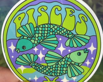 May include: A round sticker with a green, blue, and purple background. The sticker features two green fish with white stars and the word "Pisces" in green letters. The astrological symbol for Pisces is also included in the design.