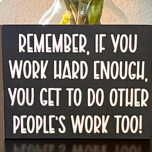 May include: A rectangular black sign with white text that reads: REMEMBER, IF YOU WORK HARD ENOUGH, YOU GET TO DO OTHER PEOPLE'S WORK TOO! The sign is positioned in front of a clear glass vase filled with vibrant red, orange, and yellow tulips.